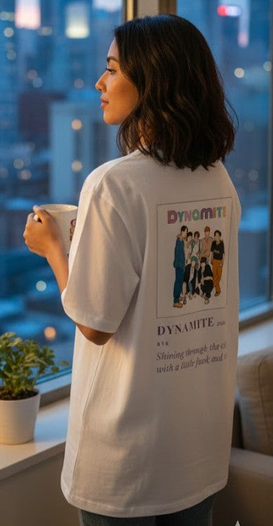 Person wearing a white t-shirt with a graphic design, standing by a window with a cityscape view.